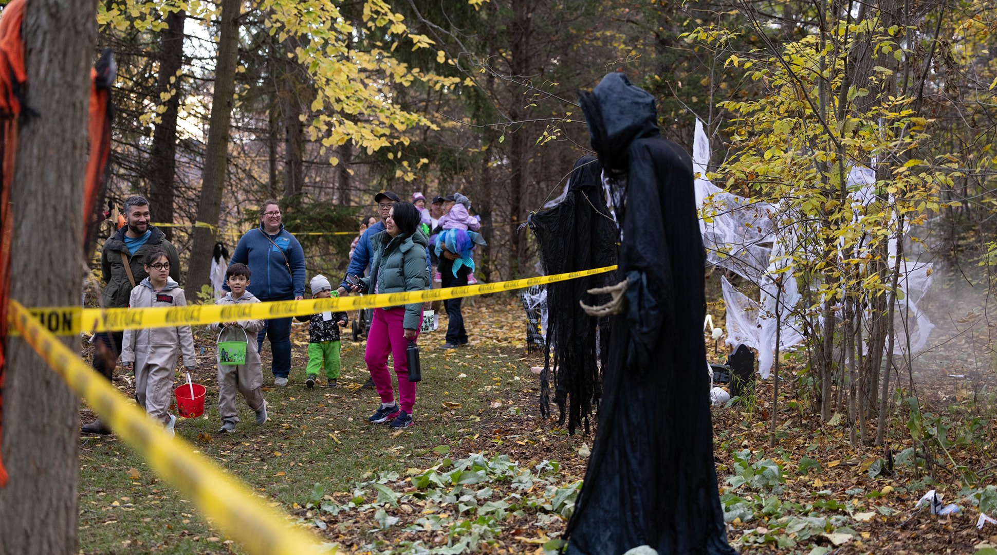Visitors walk along a wooded trail decorated as a haunted attraction during autumn. Yellow caution tape lines the path, while black-robed skeletal figures, white hanging ghost decorations, and artificial fog create a spooky atmosphere among trees with yellow fall leaves. Several children carry trick-or-treat buckets as families move through the decorated forest path.